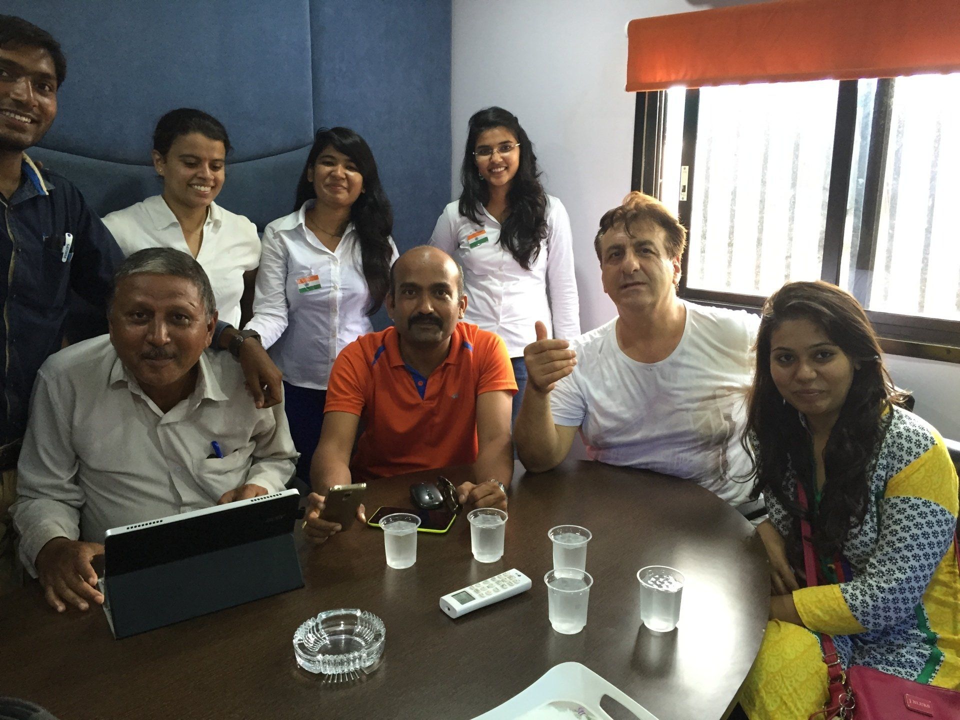 Group of people around a table, some smiling. One man gives a thumbs up. Interior office setting with window.