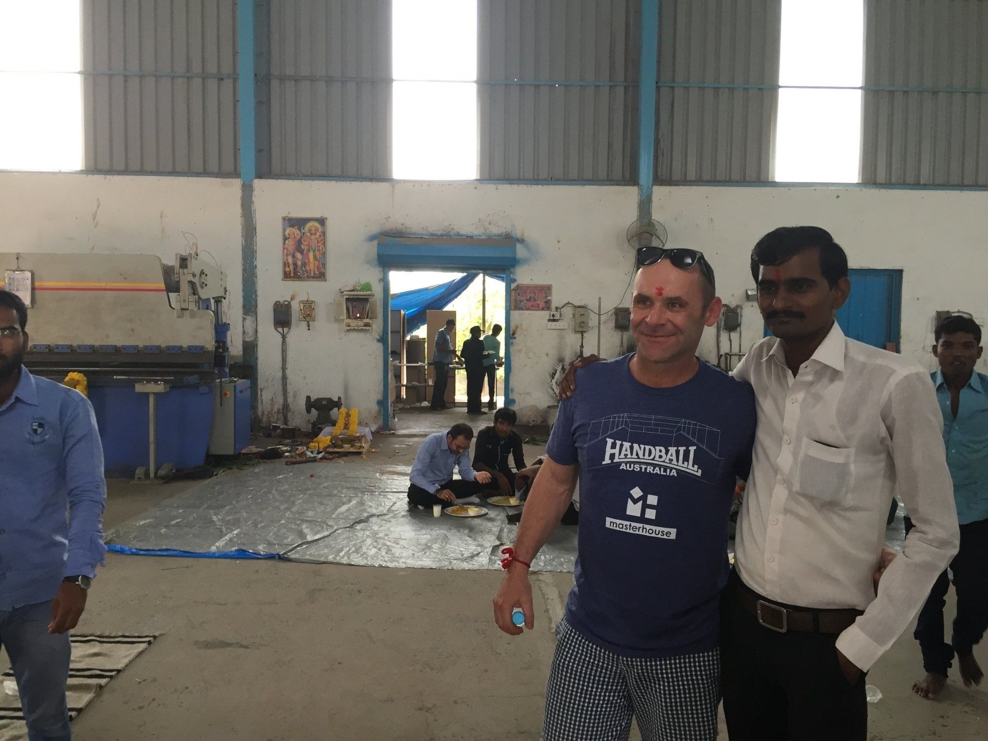Two men pose smiling inside a factory. One wears a blue t-shirt, the other a white shirt. Factory workers are visible in the background.