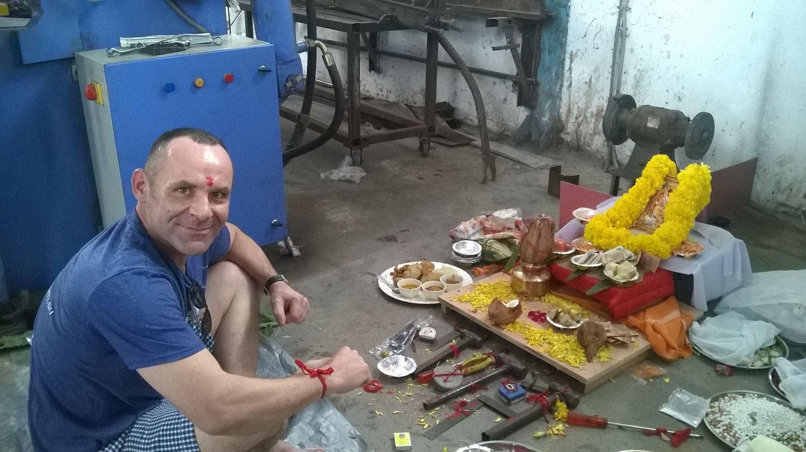 Man kneels near an altar with food, flowers, and tools in a workshop.