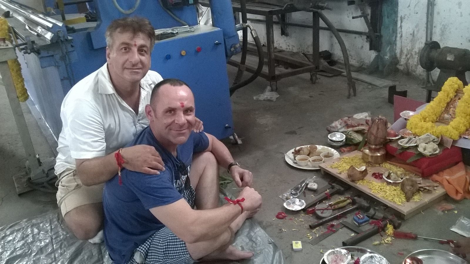 Two men pose near an industrial machine and a religious offering. One man has a red mark on his forehead, and both have red wristbands.