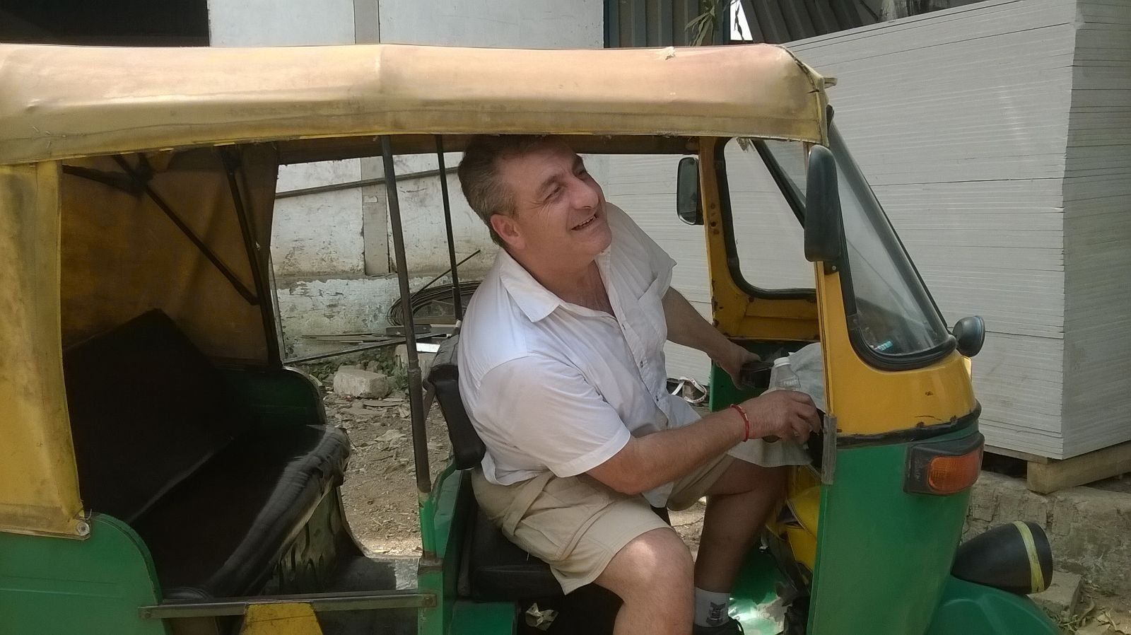 Man driving a green and yellow auto-rickshaw, smiling, in an outdoor setting.