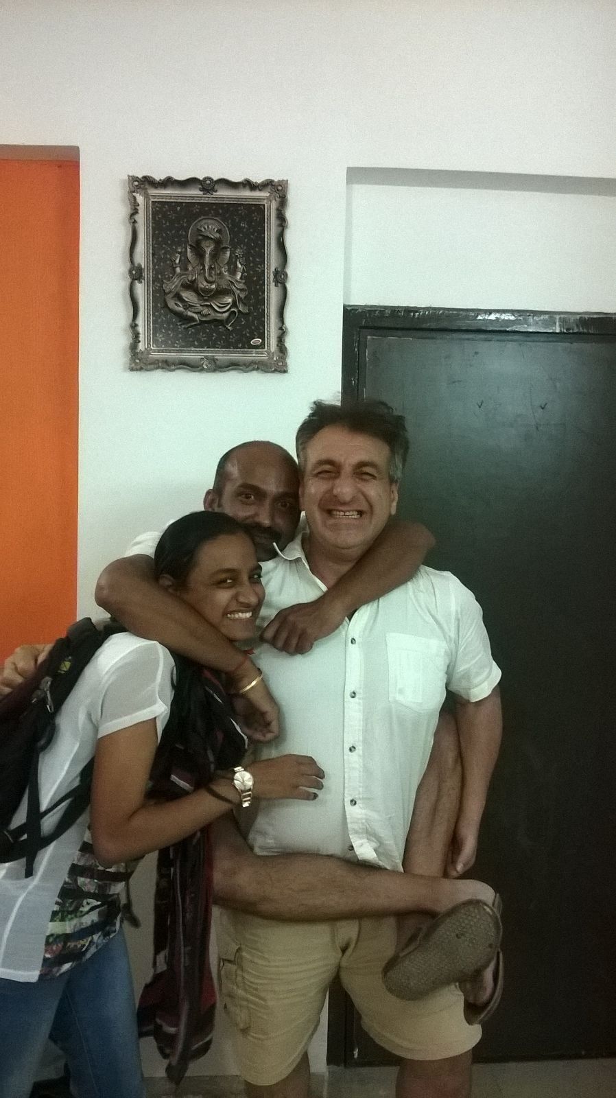 Three people smiling, hugging indoors. Man in shorts and shirt is the focus, flanked by two others. A wall with a framed art piece and an orange wall are visible.