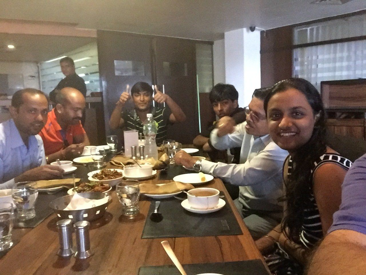 Group of people at a restaurant table, eating and smiling. Bottles, plates, and glasses visible.