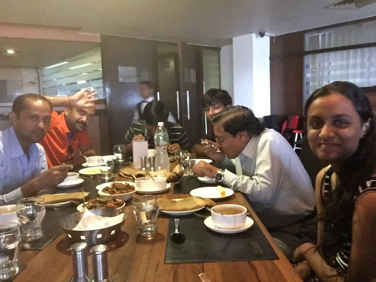 People seated around a wooden table in a restaurant, eating. Various food dishes and glasses are on the table.