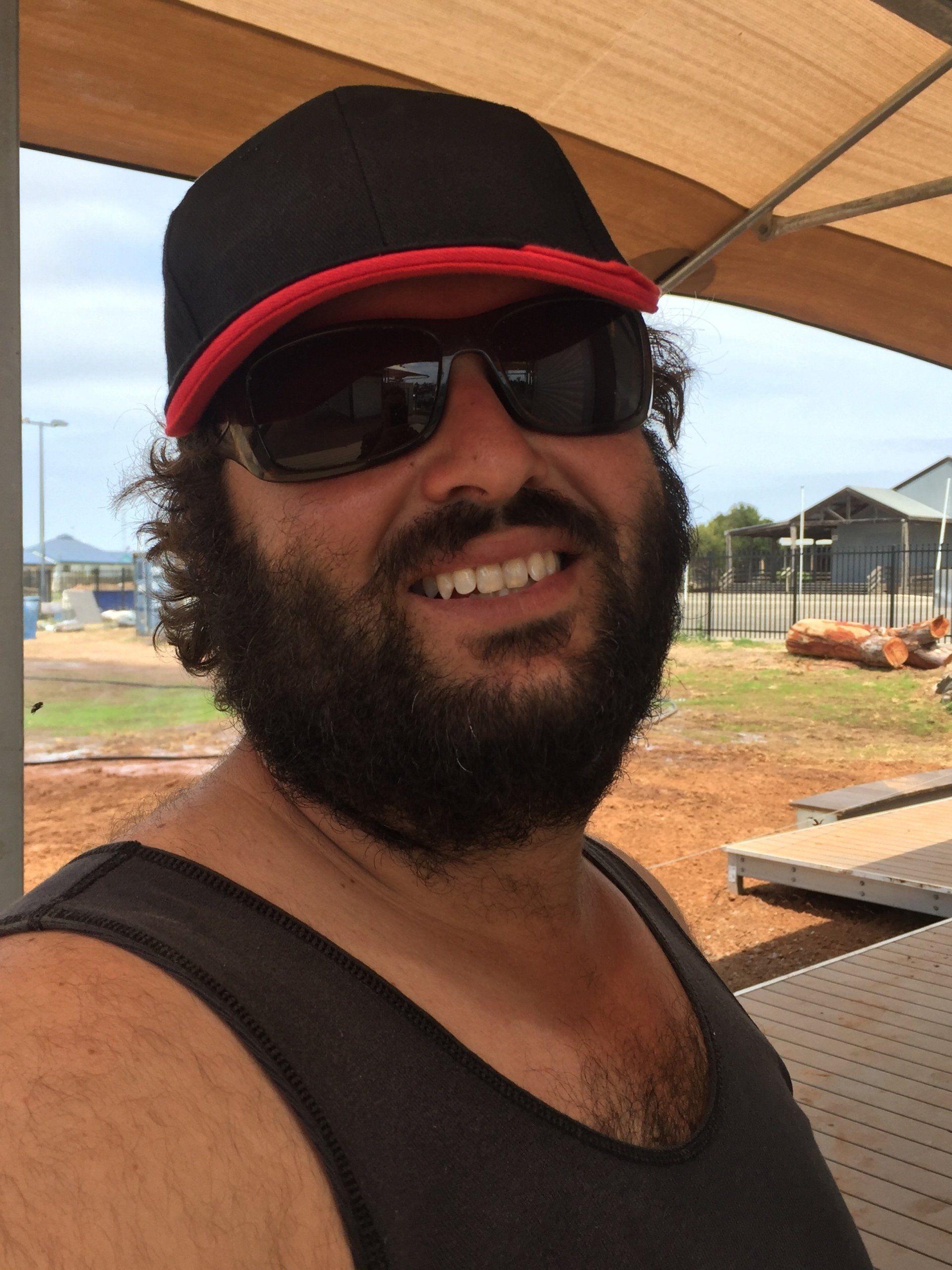 Man with a beard and sunglasses, wearing a black cap with a red brim, smiling outdoors.