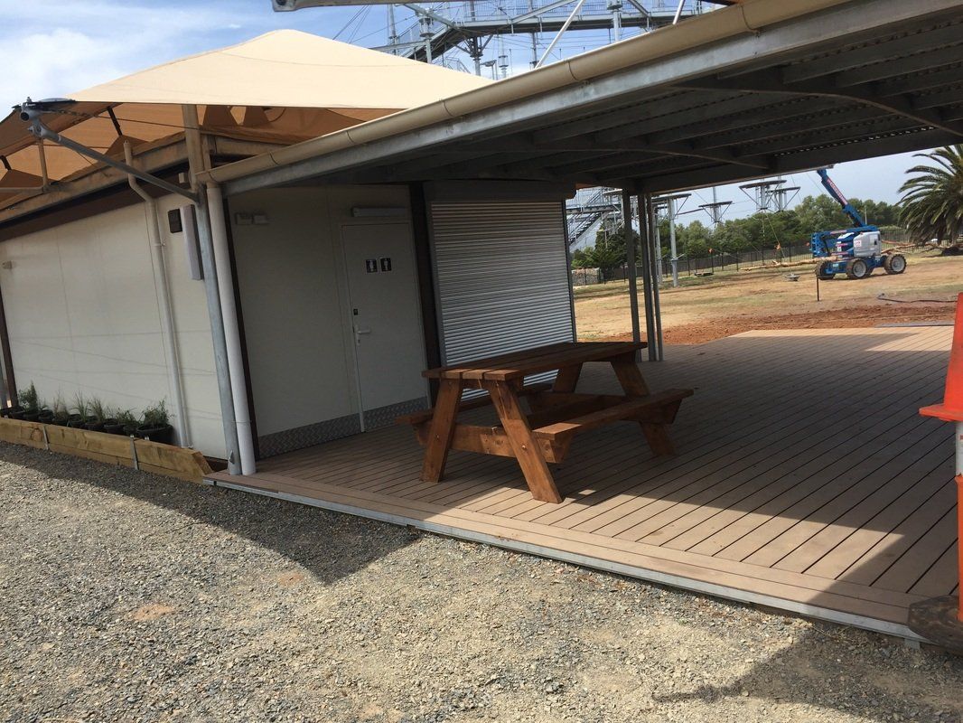 A picnic table under a covered area, next to a building with a sunshade, outdoors.