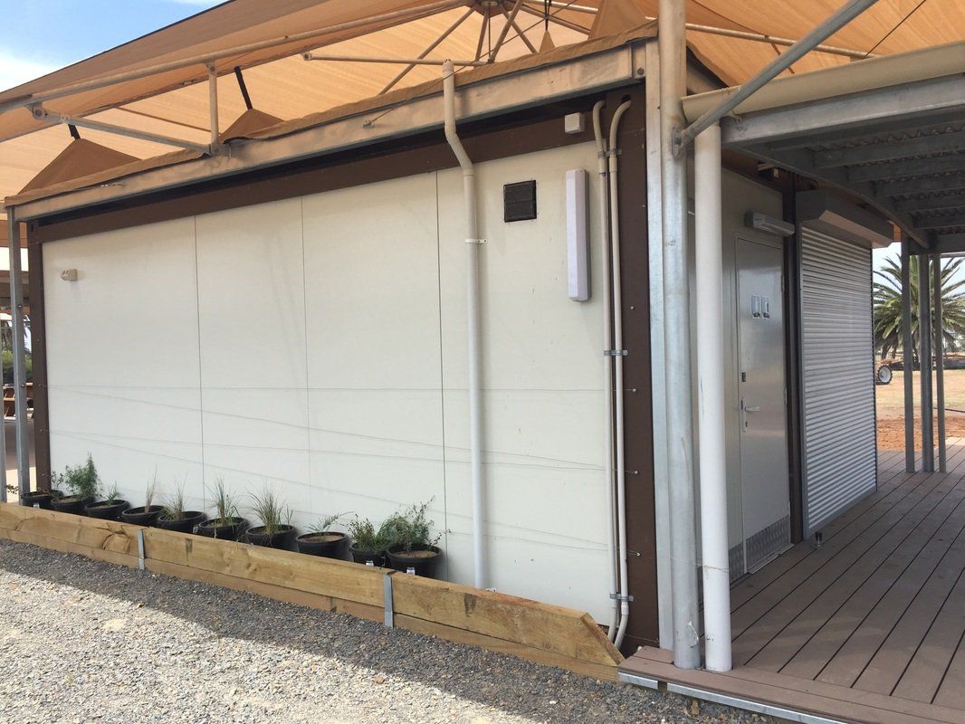 White building with brown trim under a shade structure, pots of plants along the base, gravel ground.