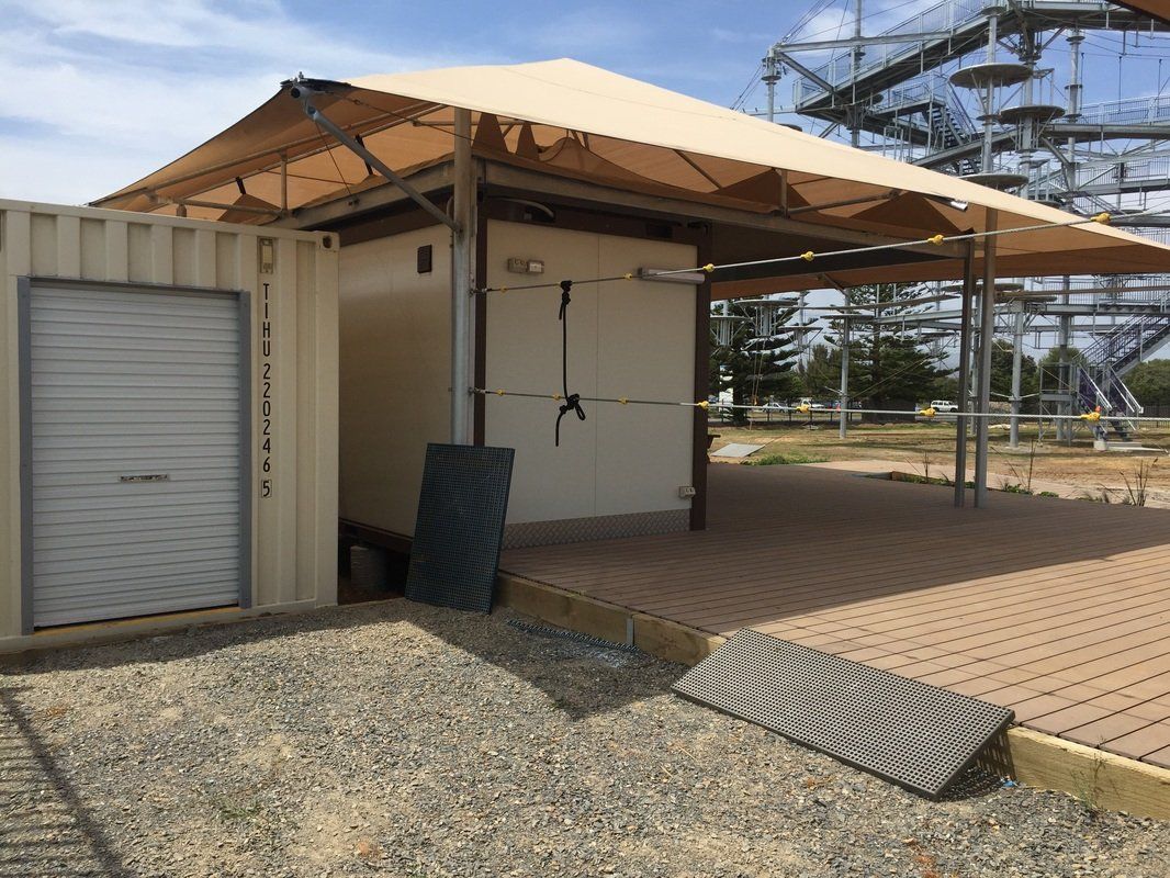 Shipping container office with sunshade, on a brick patio, near an outdoor adventure course.