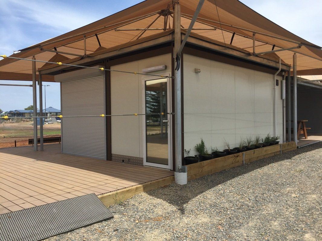 Small white building with a deck, tan shade roof, and gravel ground.