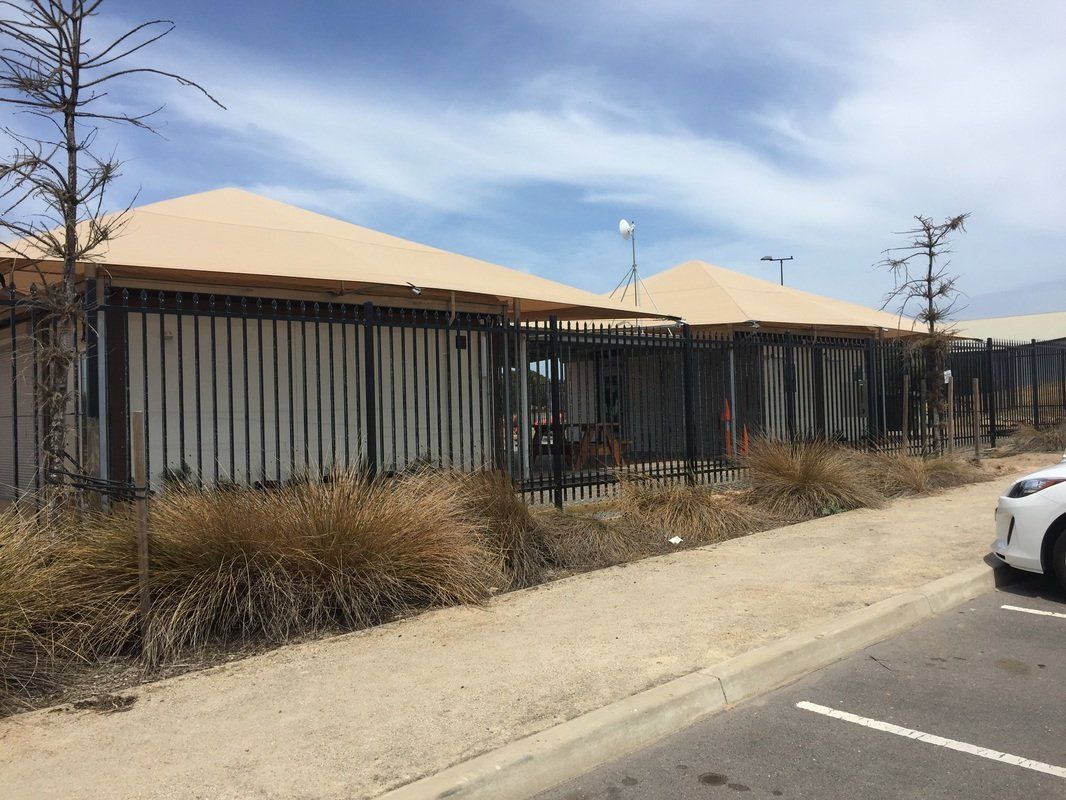Buildings with beige roofs and vertical slat walls, next to a sandy sidewalk and dry bushes. Cloudy sky.