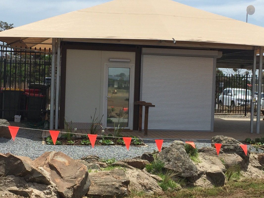 Small beige-roofed building with corrugated metal siding, glass door, and orange safety flags in front.