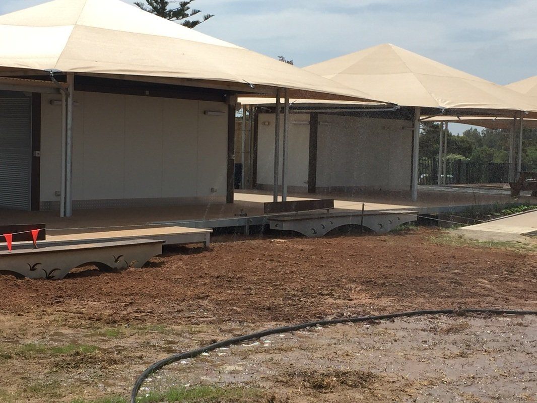 Several tan-roofed buildings with open sides, set on a raised platform, surrounded by dirt and grass.