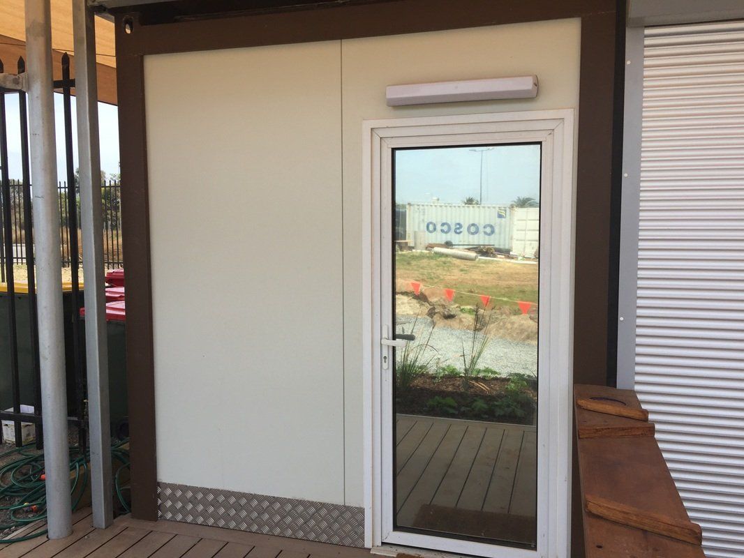 Exterior of a small white modular building with a glass door, brown trim, and a small ramp.