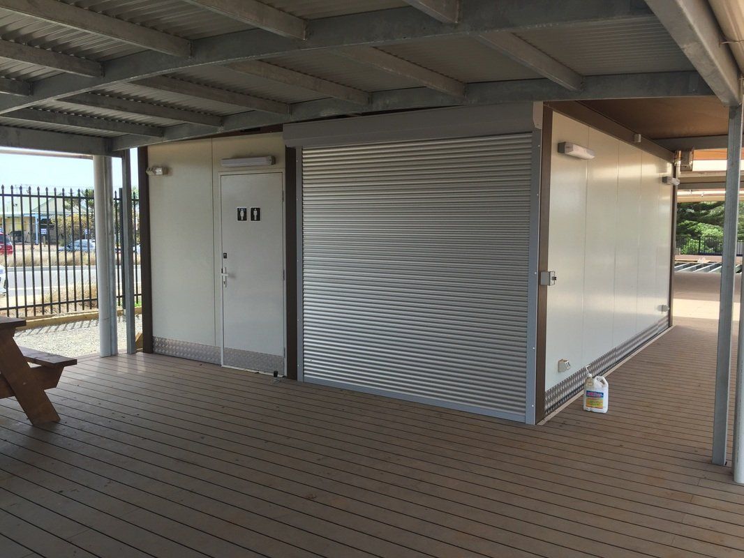Public restroom building under a covered deck. One door has a restroom symbol. A roll-up door is closed.