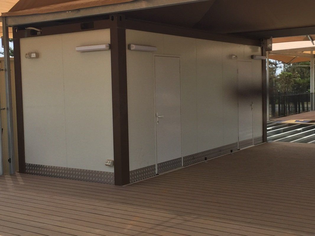 White rectangular building with brown trim, door, under a shaded canopy.