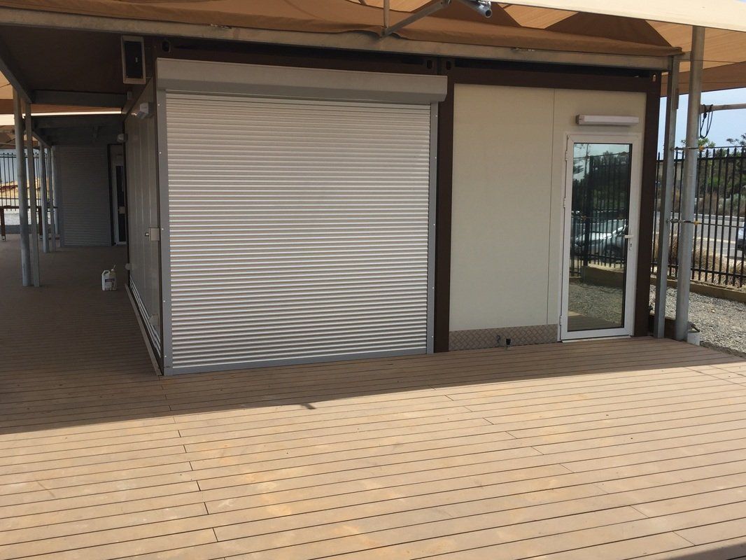 A small building with a closed roller door and a side glass door under a shaded canopy.