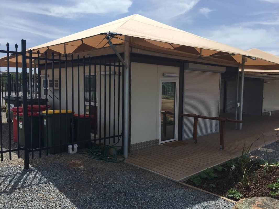 Tan awning covers small building with black fence and wooden deck. Gravel path, trash cans present.