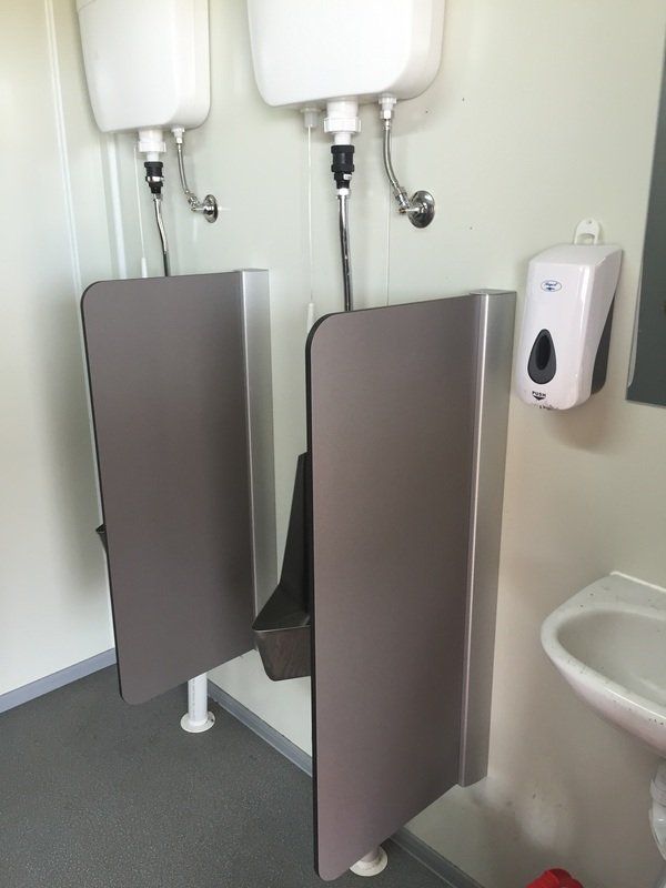 Two urinals with dividers in a public restroom. White walls, grey dividers, and a soap dispenser are visible.