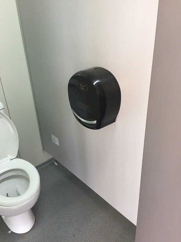 Black hand towel dispenser on a light-colored wall, next to a toilet in a bathroom.