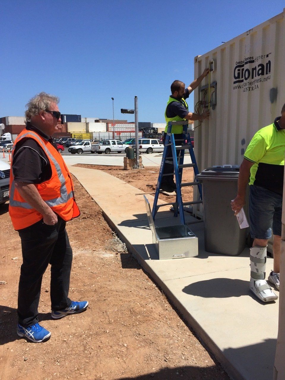 Three workers by a storage container on a sunny day; one on a ladder, two on the ground.