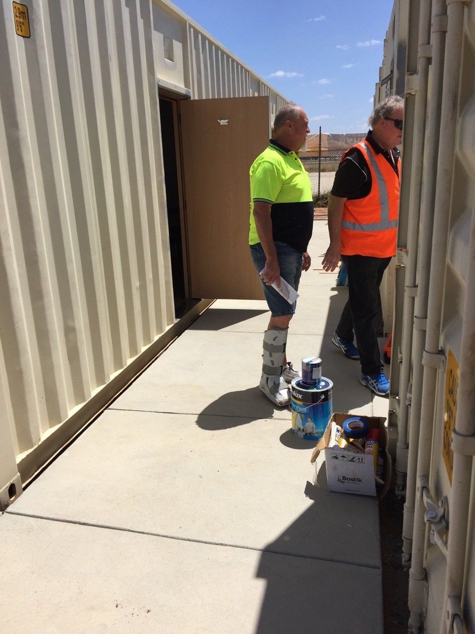 Two men stand in a narrow space between shipping containers. One wears a cast on his leg.