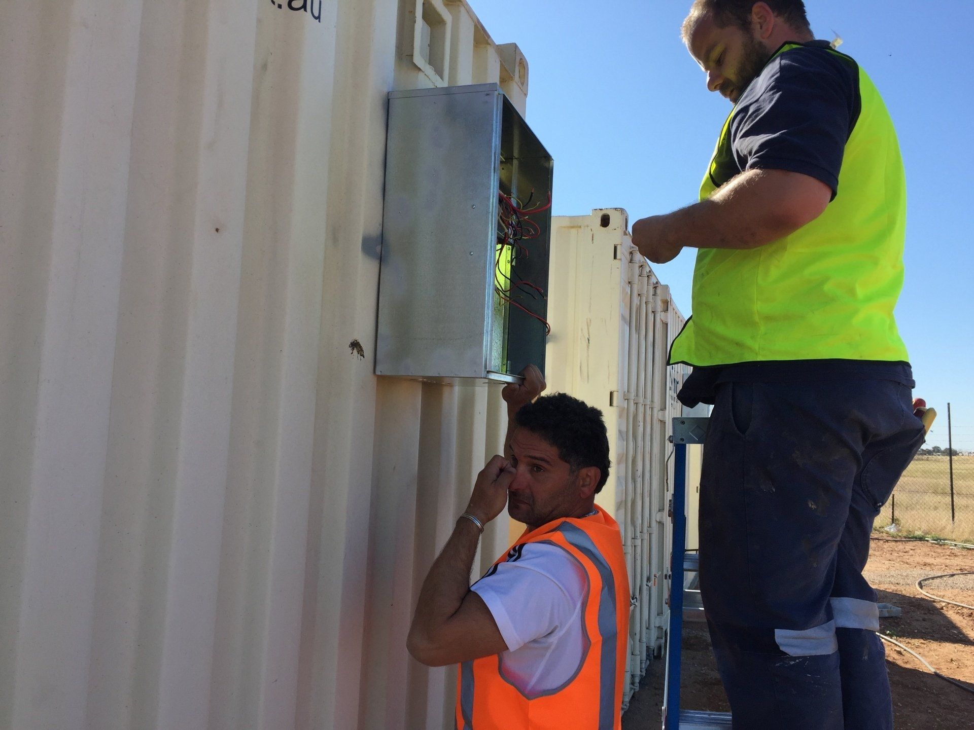 Two workers in safety vests installing something on a beige building in a sunny, outdoor setting.
