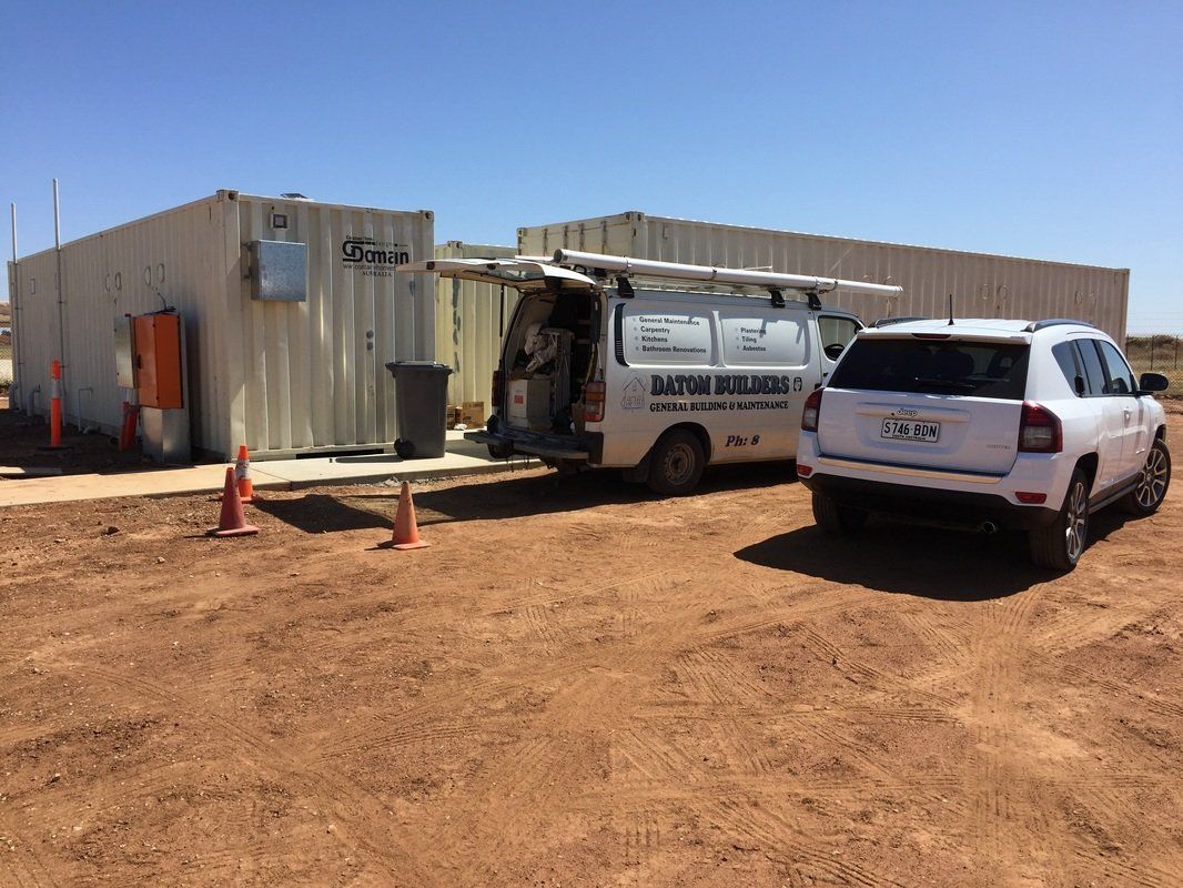 A white van with ladder, a white SUV, and two rectangular structures are parked on dirt ground under a bright blue sky.