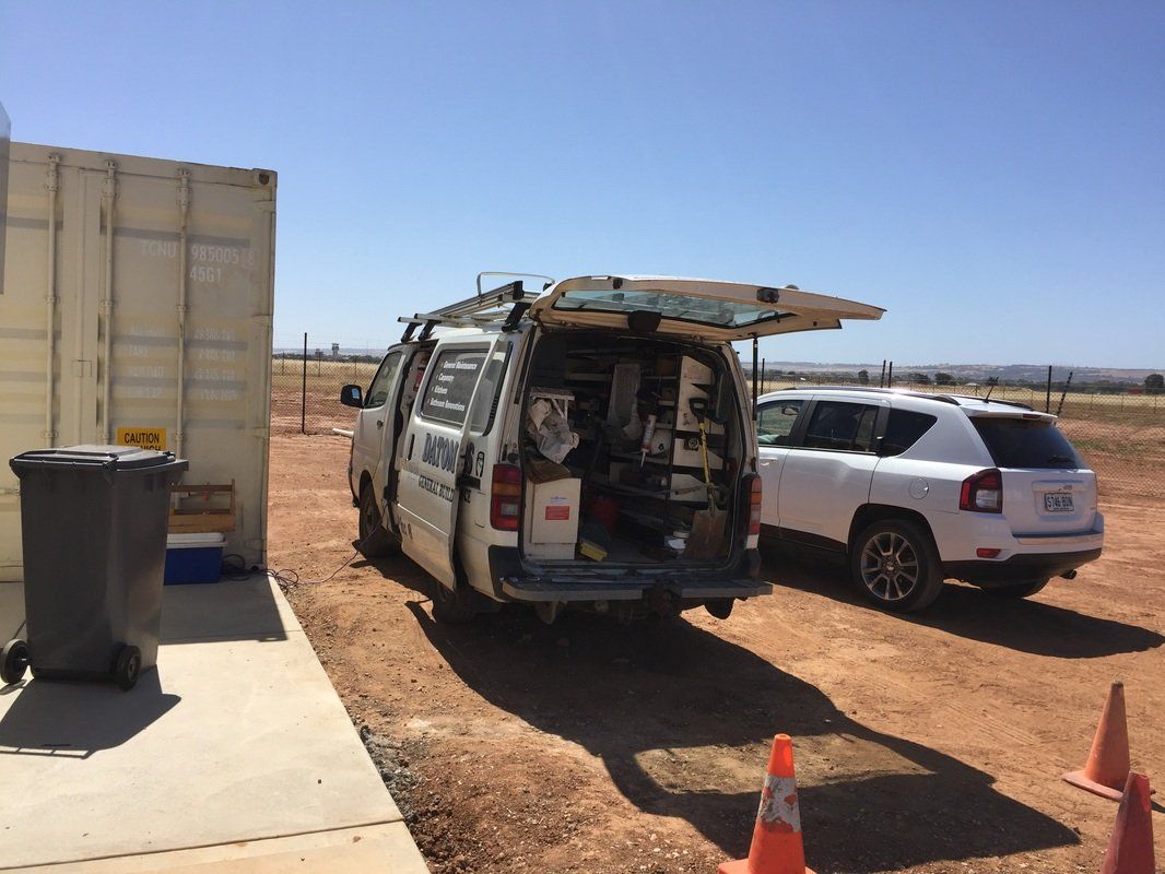 A work van parked near a storage container with its back door open. White SUV beside it, blue sky.