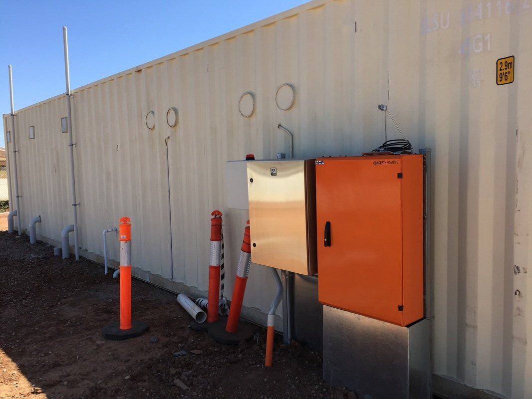 Orange electrical box attached to a corrugated white wall with orange traffic cones.