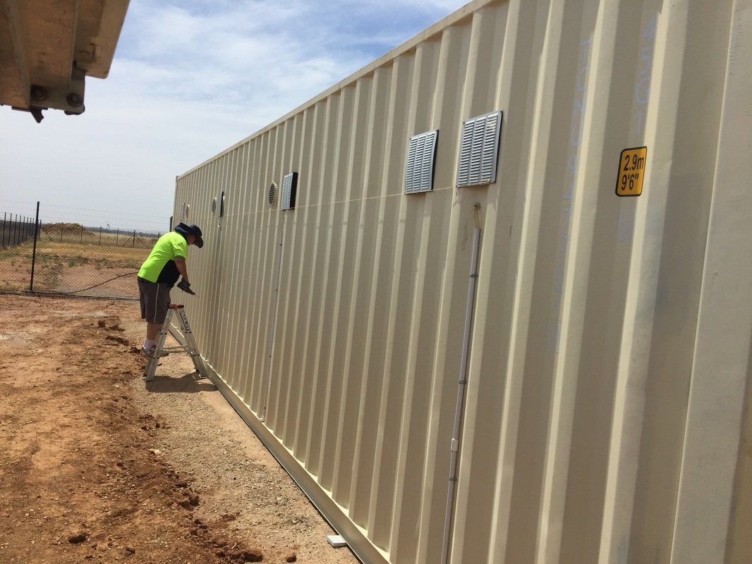 A person in a neon green shirt works on the side of a beige shipping container outdoors.