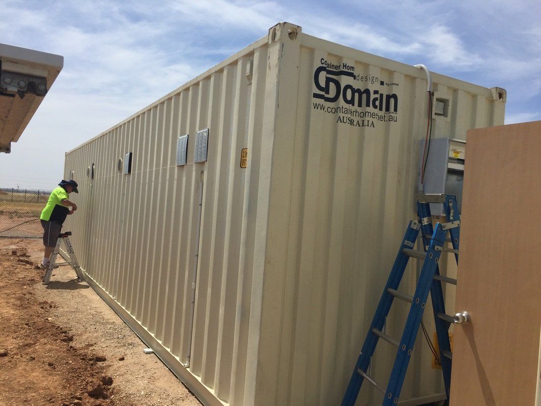 A tan shipping container with vents, a worker, and a ladder, outdoors on a construction site.