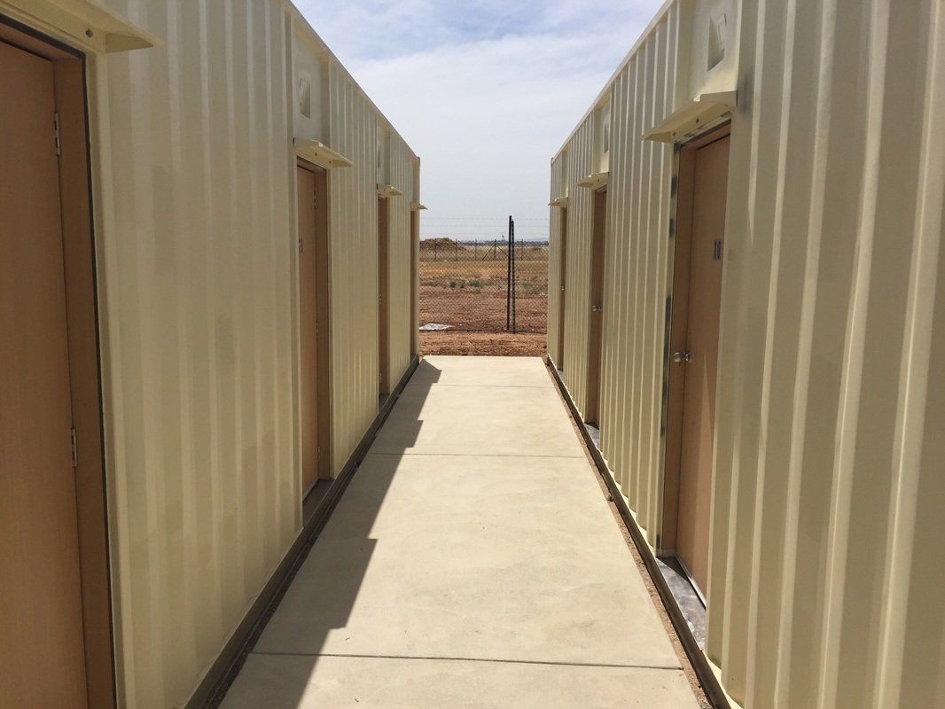 Two tan corrugated metal buildings with brown doors face each other, creating a concrete walkway.