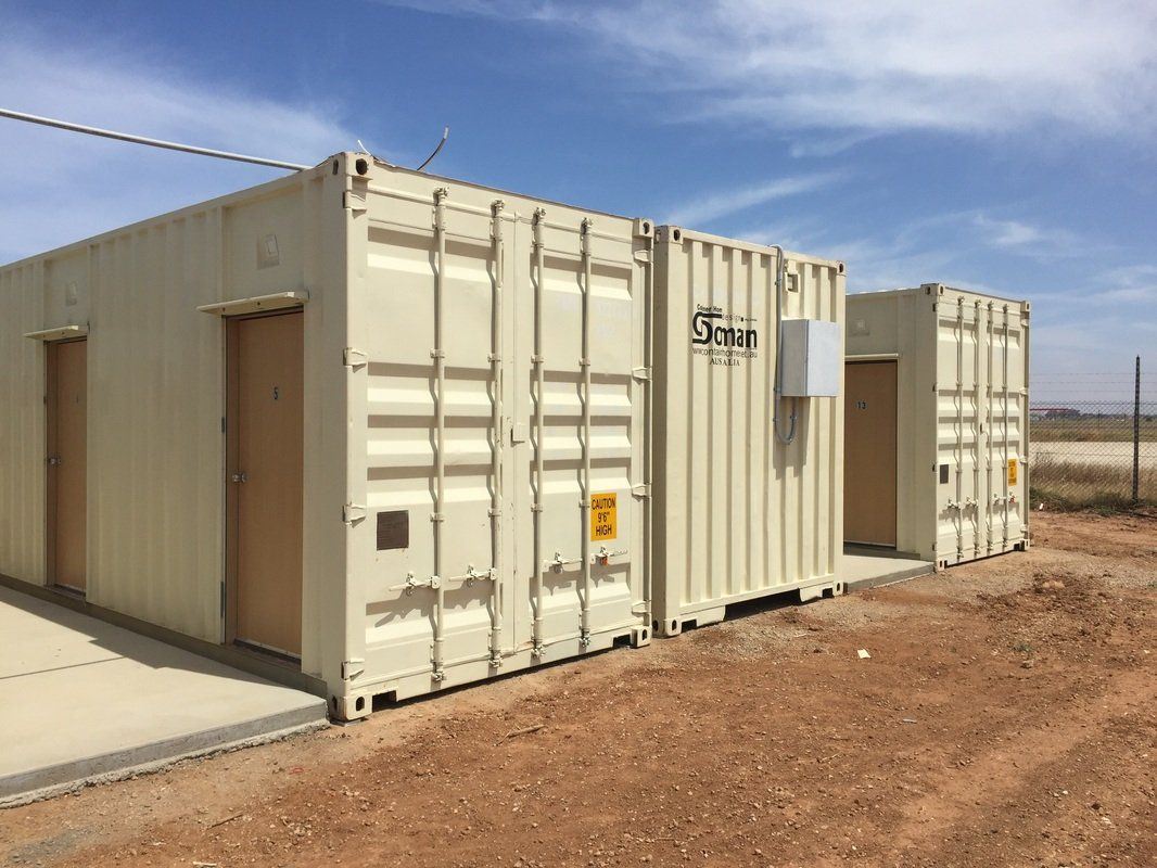 Two beige shipping containers repurposed as buildings, on a dirt lot, with brown doors and a blue sky.