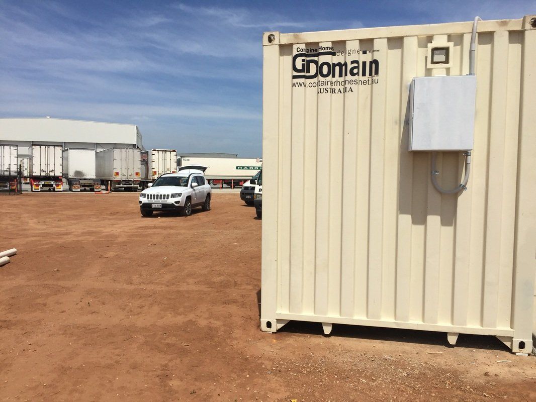 Beige shipping container with antenna, parked vehicles, and industrial setting.
