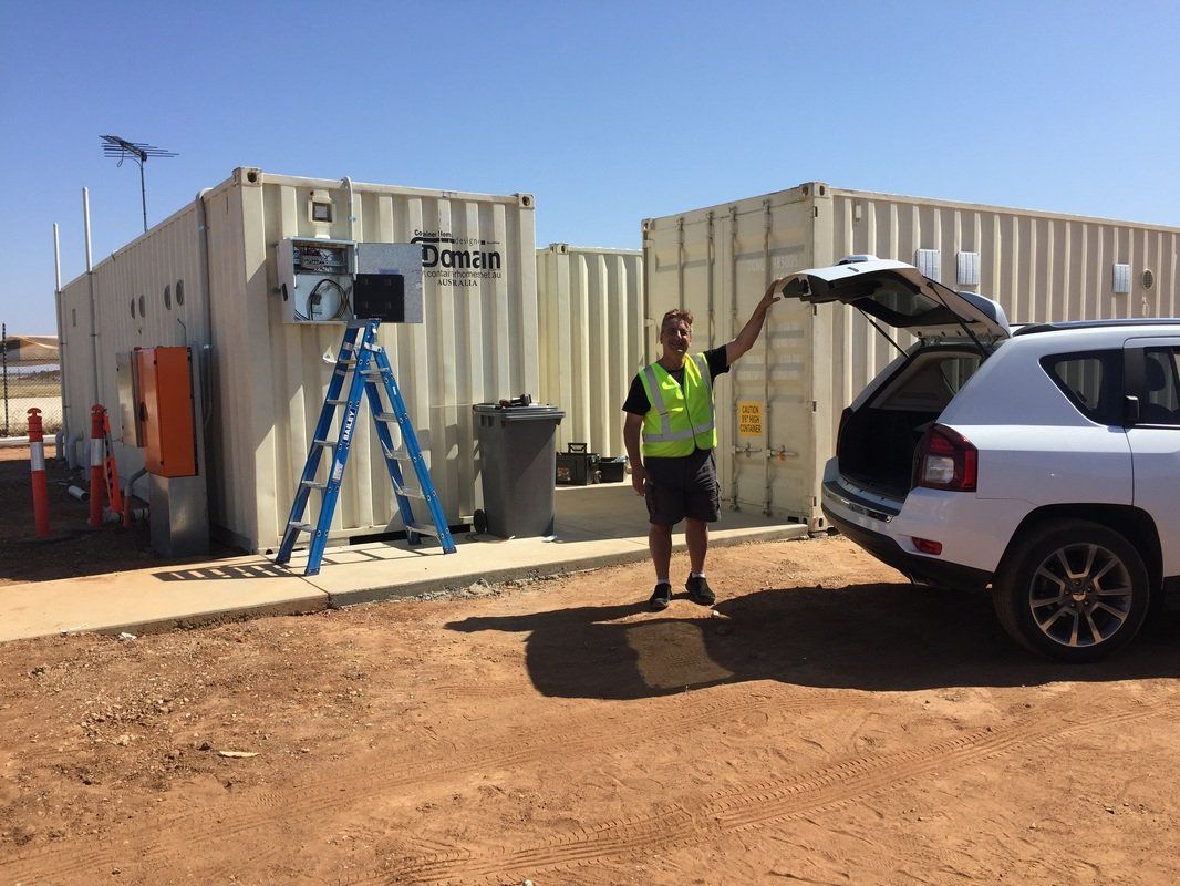 Man in high-vis vest near shipping containers, gesturing toward a car with an open hatch. Outdoor setting, sunny.