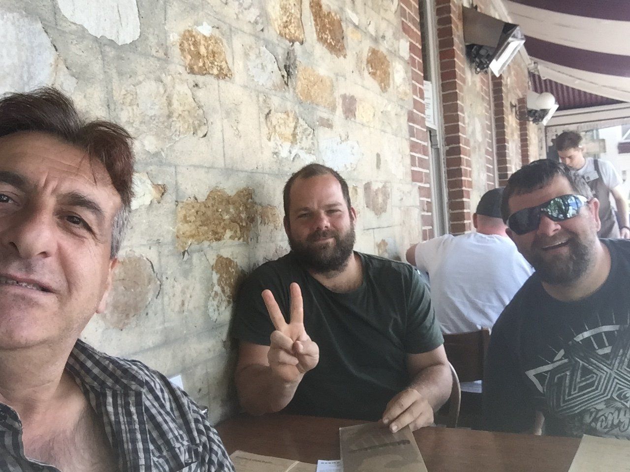 Three men at a table outside, one giving a peace sign. Stone wall background, sunny day.