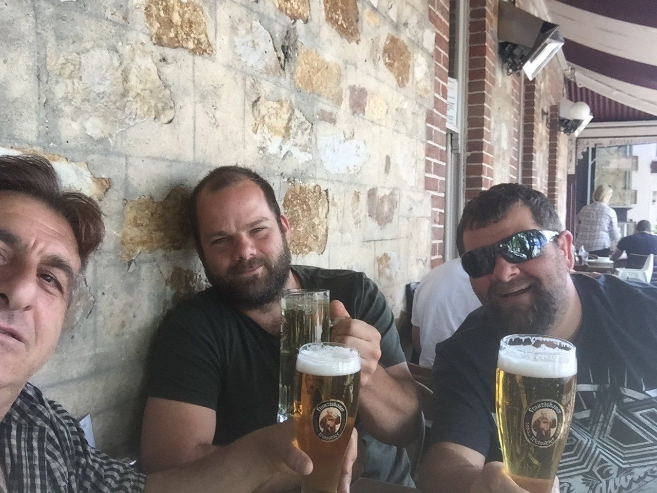 Three men with beers, toasting, outdoors near a stone wall. One wears sunglasses.