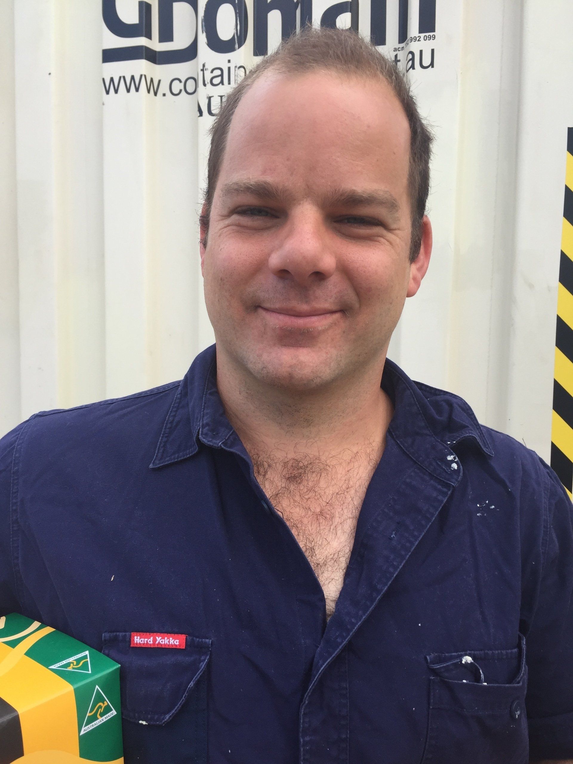 Man in blue work shirt smiles, holding a yellow and green object, in front of a container.