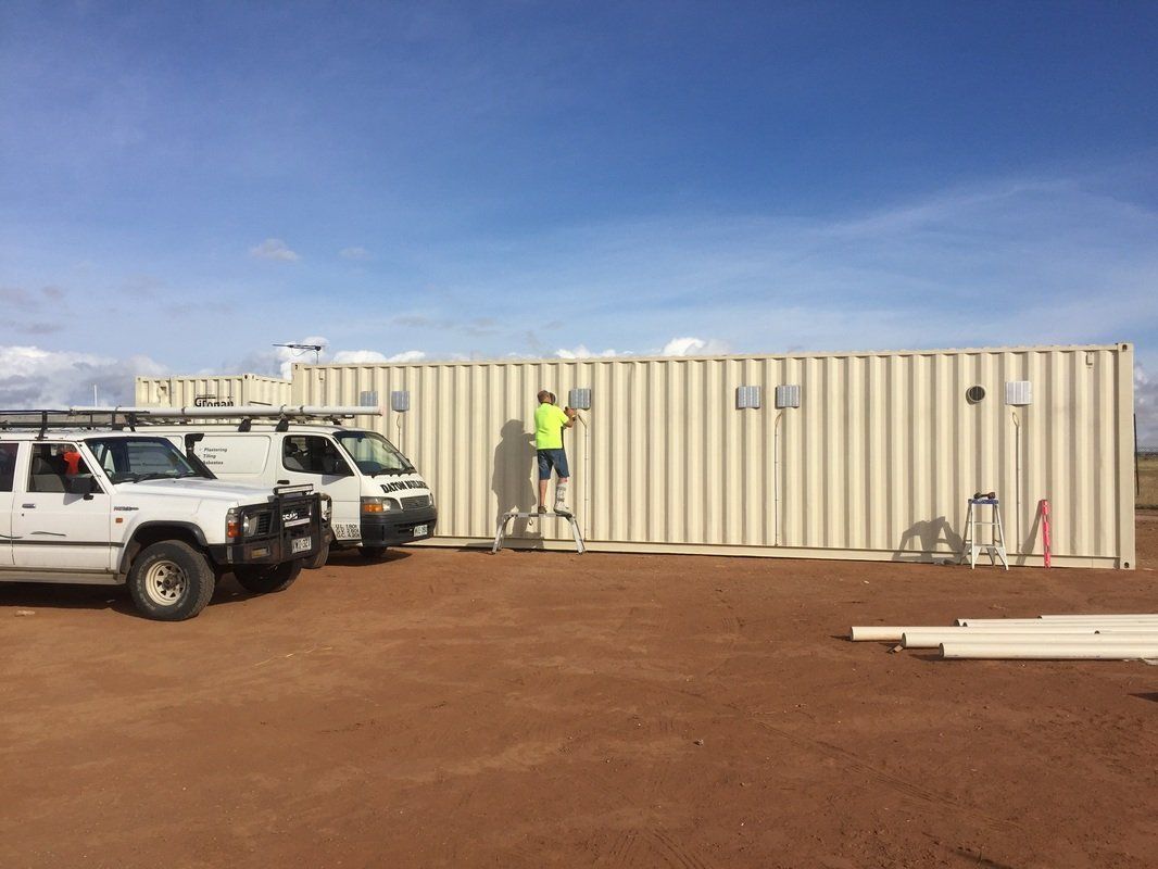 White work trucks parked by light tan shipping containers; person on ladder working on the containers.