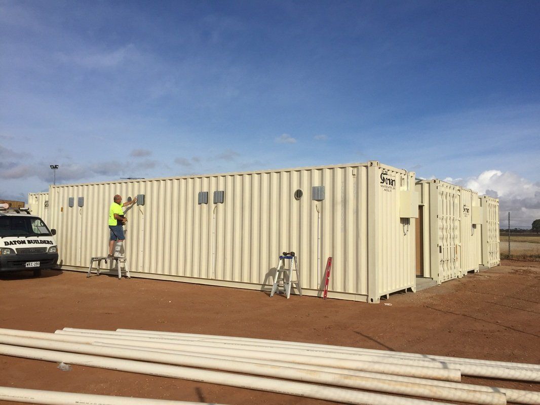 A man on a stepladder works on a structure made from shipping containers, with a truck nearby and PVC pipes in the foreground.