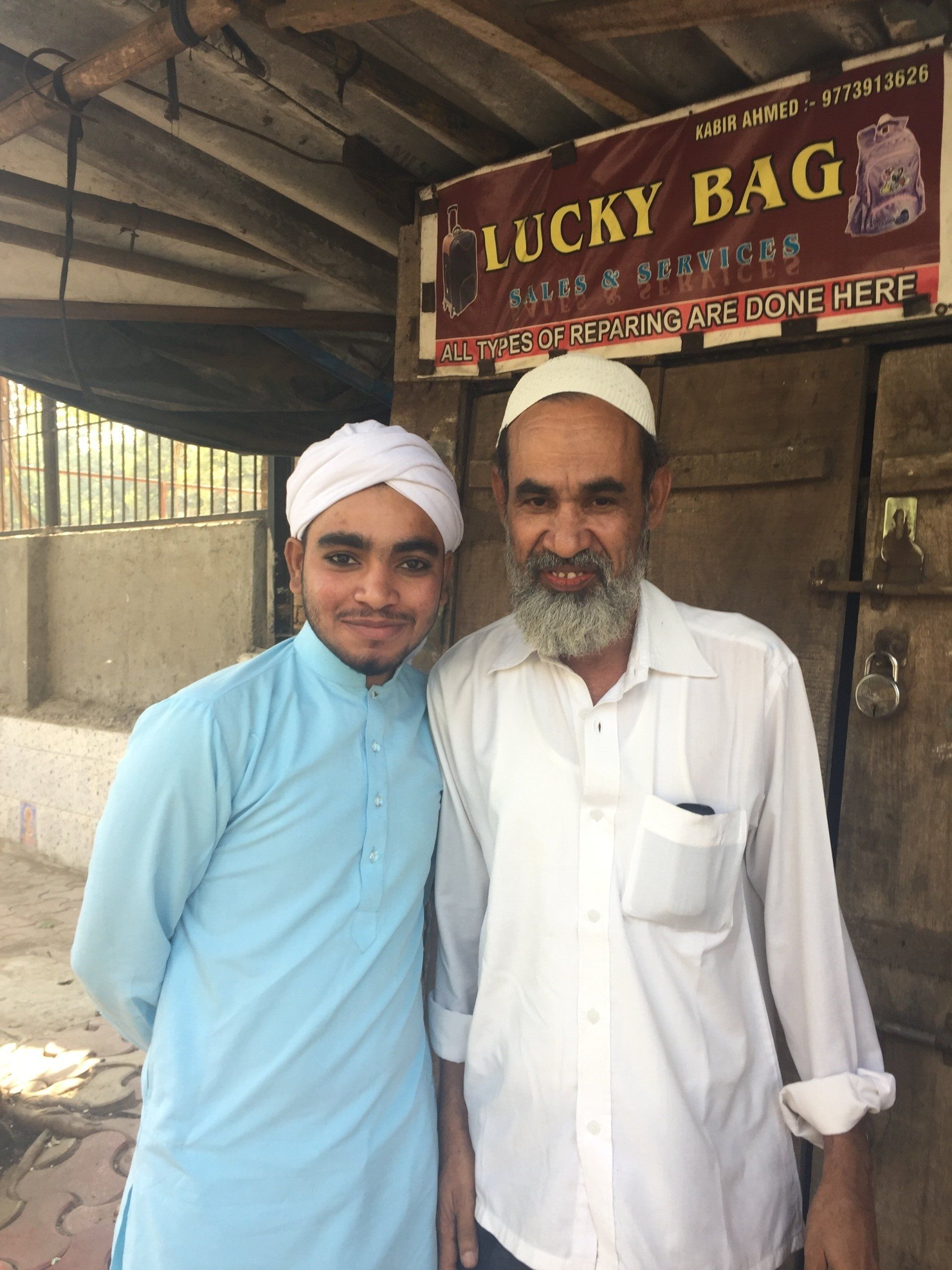 Two men stand outside a shop. One wears a blue tunic and turban, the other a white shirt. A