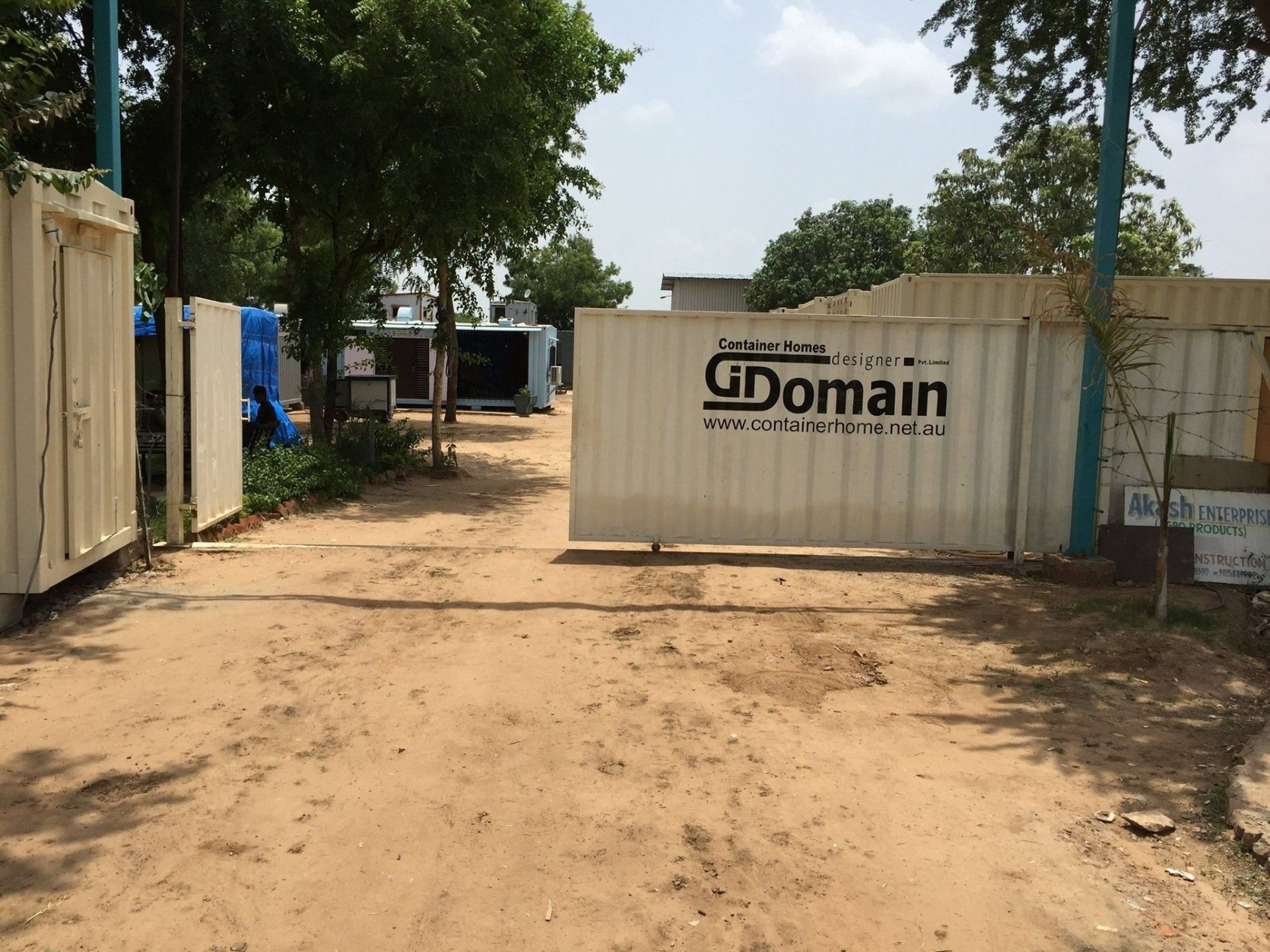 Entrance to a facility with a large white gate, on a dirt road, with trees and buildings in the background.