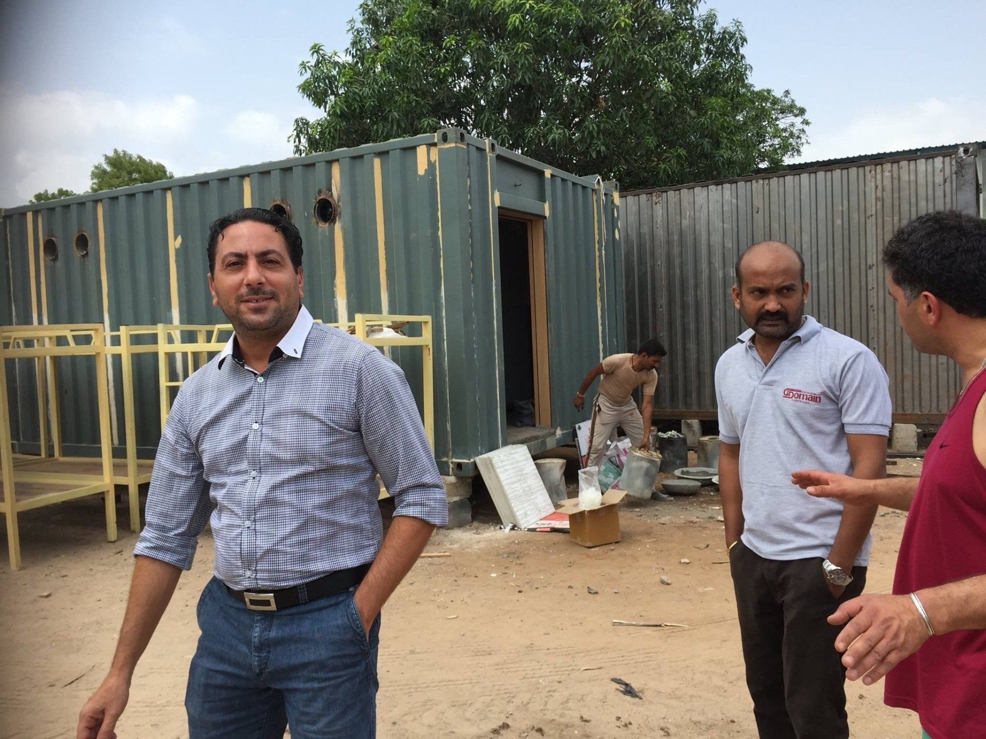 Three men stand outside shipping container structures. One wears a patterned shirt, others wear casual tops. Outdoor setting.