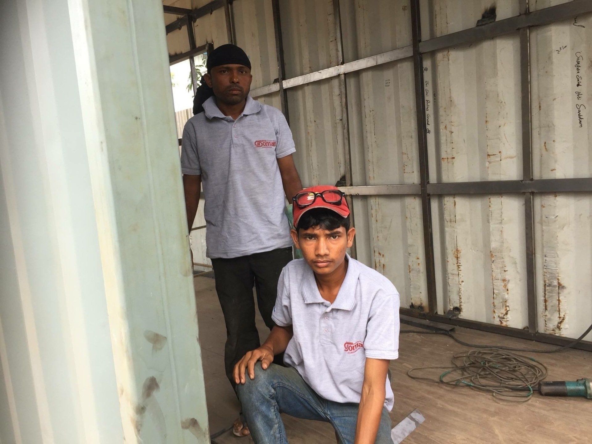 Two workers in matching gray shirts in a construction setting; one kneels, the other stands behind.