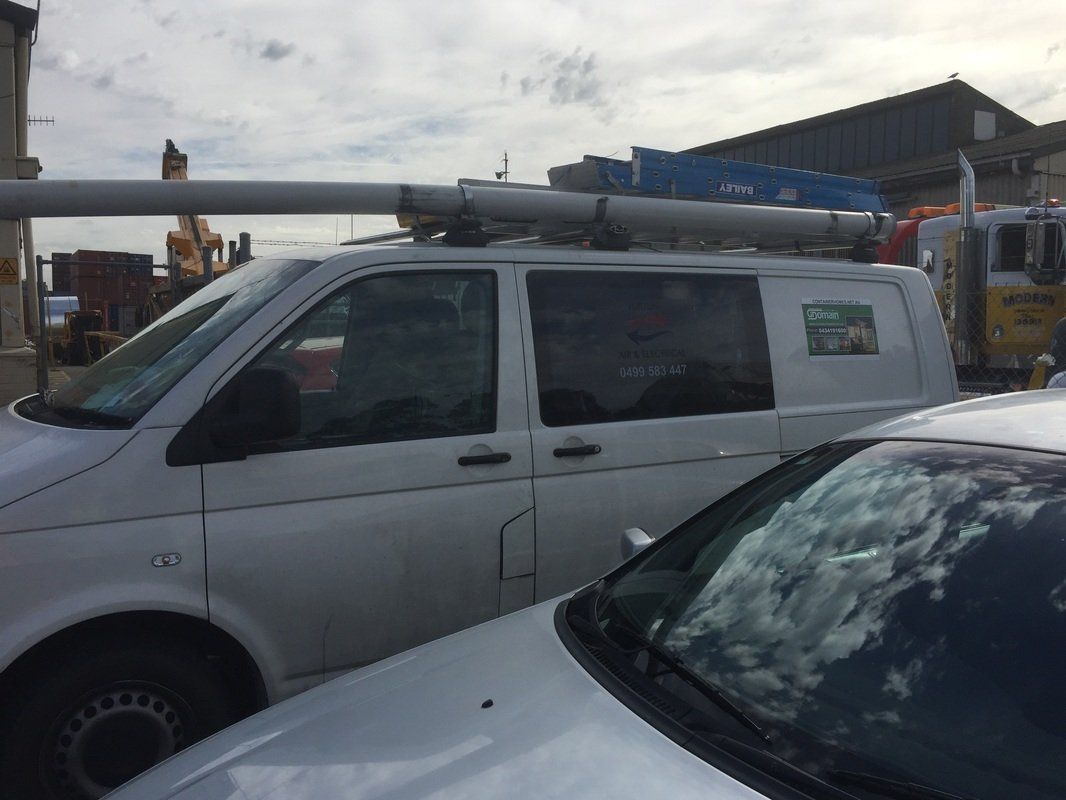 White van with equipment on the roof, parked next to a silver car; buildings and sky in background.