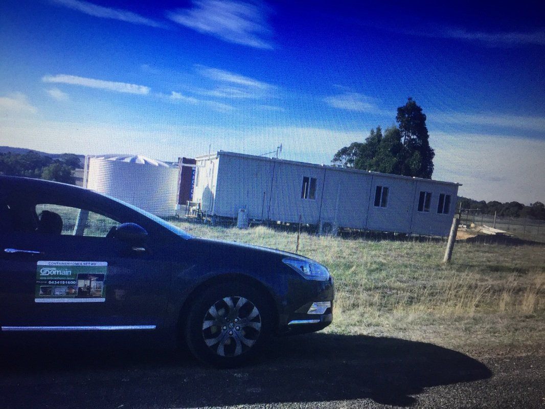 Dark car parked in front of a white building with silos under a blue sky, in a grassy field.