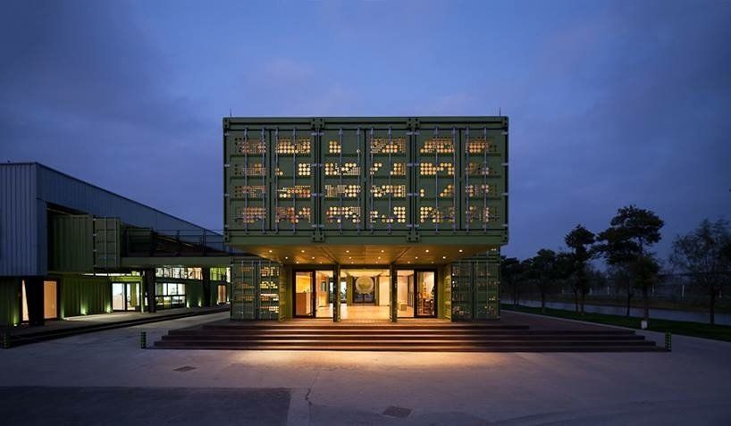 Green shipping container building with lit windows, at dusk.