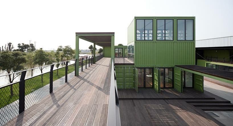 Green shipping container building with wooden walkway and large windows.