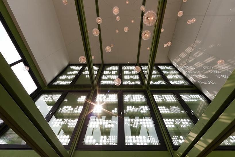 Looking up at a glass ceiling with sunlight, surrounded by green beams and floating light fixtures.