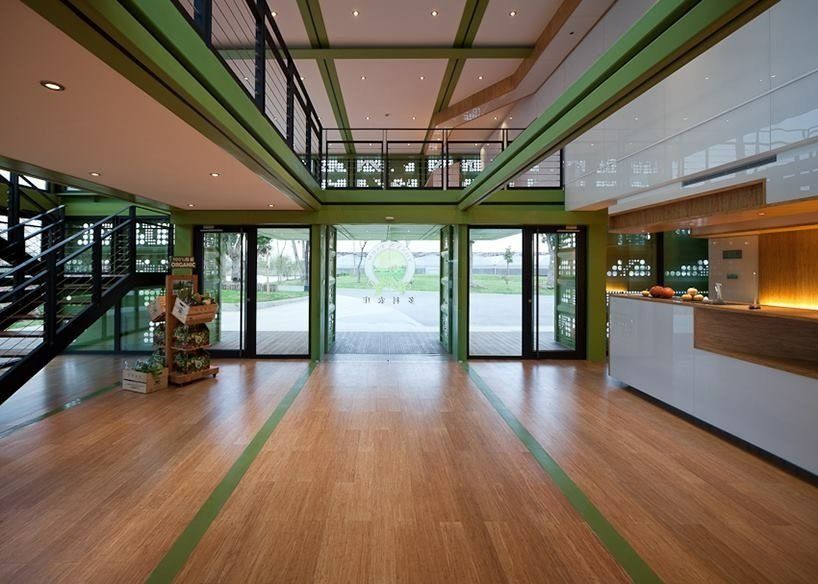 Interior view of a modern building with large glass doors and wooden floors, accented by green trim and a second-floor balcony.